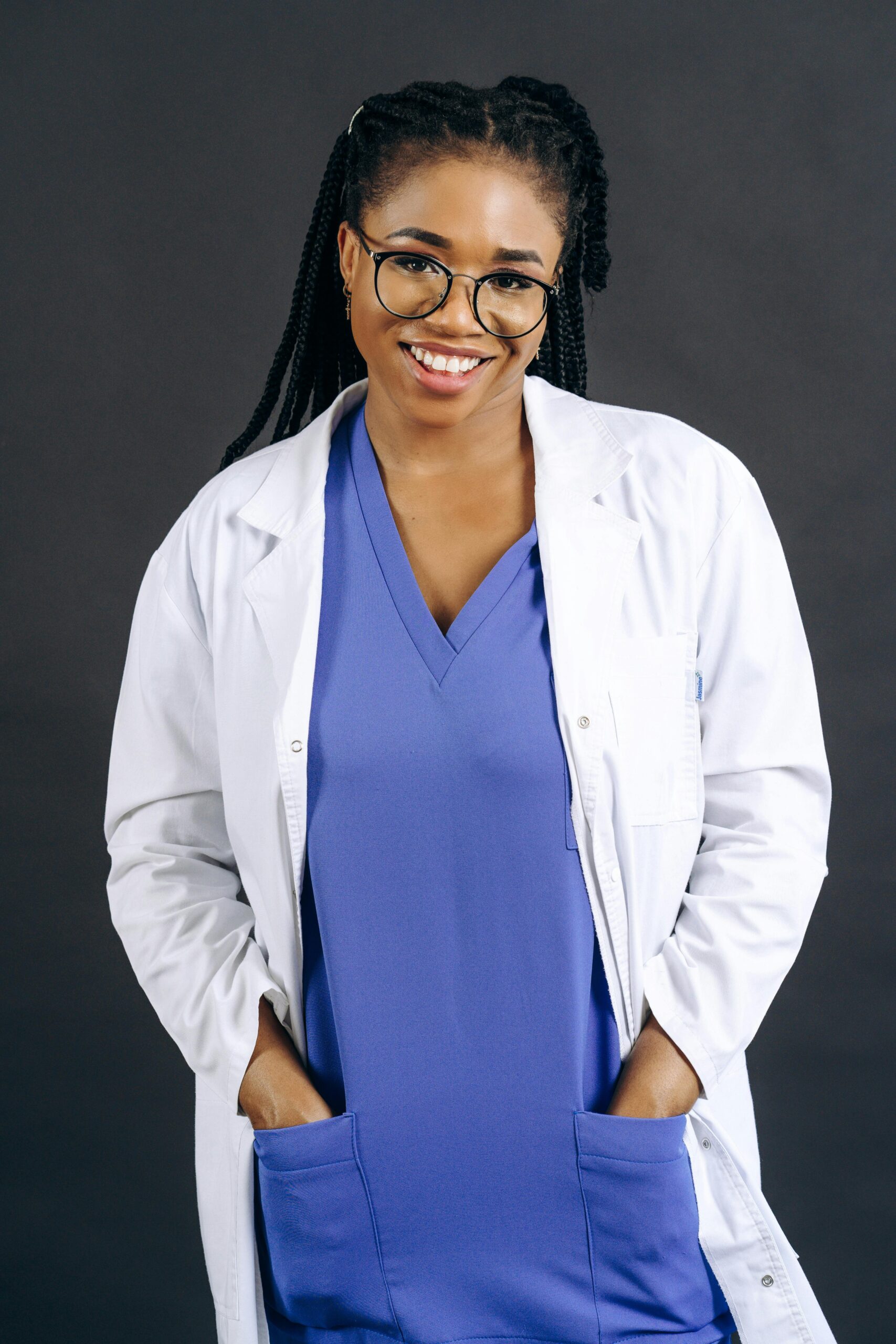 Smiling black woman doctor in scrubs standing confidently with hands in pockets.