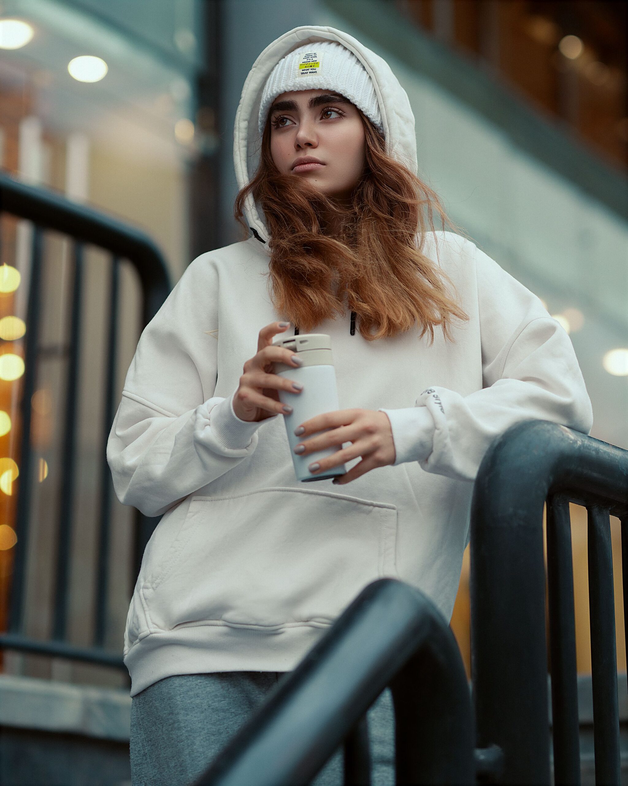 Portrait of a woman in a white hoodie holding a thermos in an urban setting.