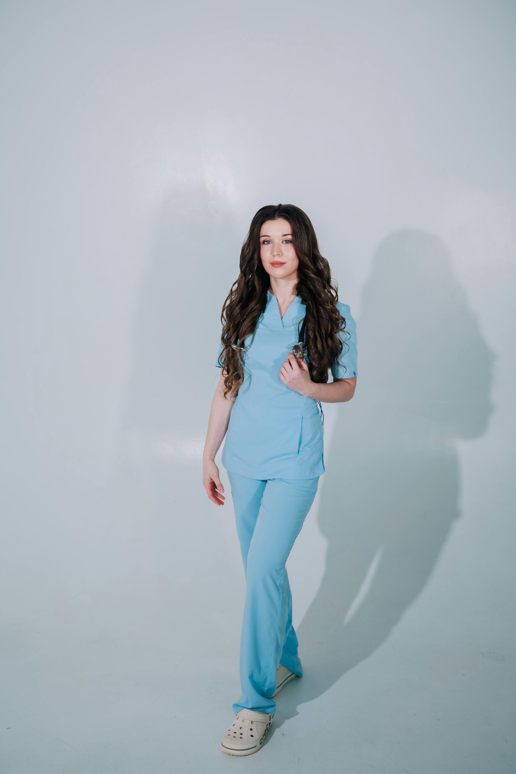 Confident female medical professional wearing blue scrubs in a bright studio setting.