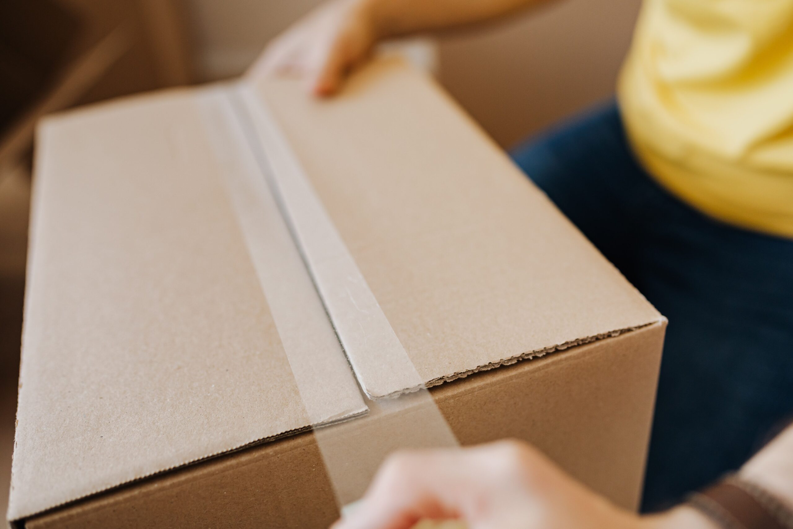 Crop anonymous courier in yellow shirt packing purchase and sealing carton box with tape before deliver