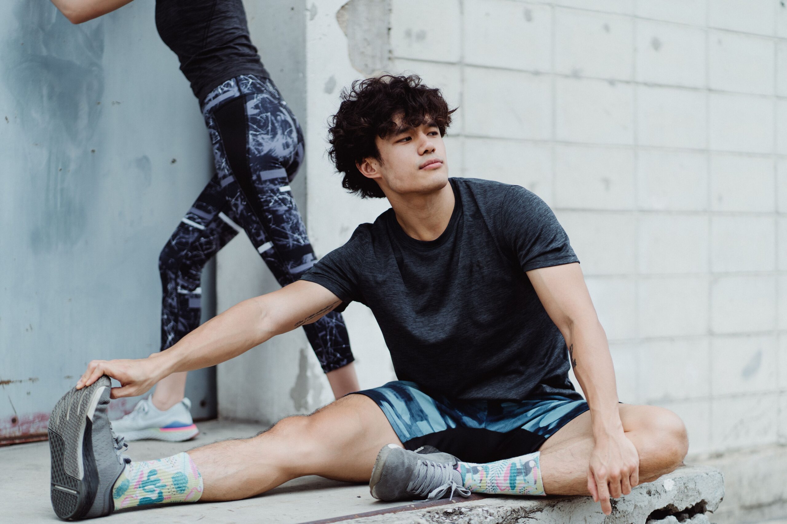A young man sitting on concrete, stretching before a workout, with another person in the background outdoors.