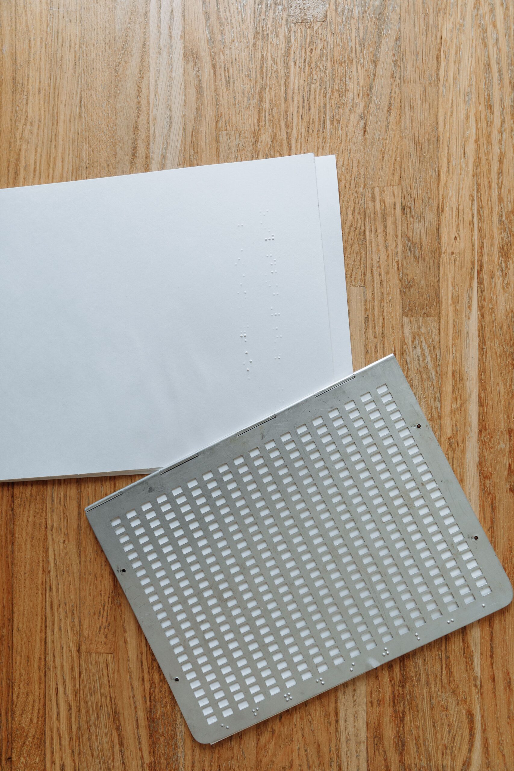 Braille paper with a metal guide indicating a tactile writing system on a wooden surface.