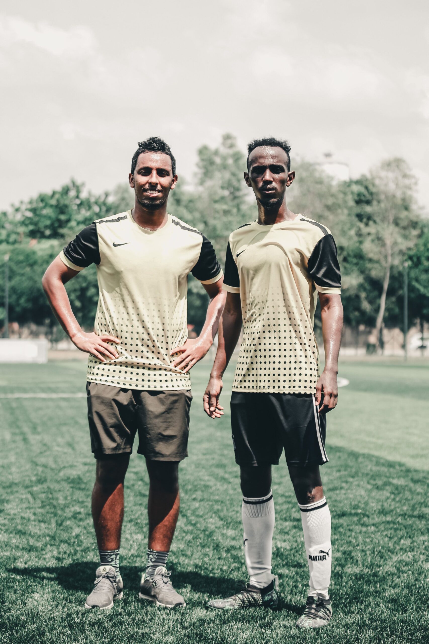 Two soccer players in uniforms pose confidently on a grassy field during the day, showcasing teamwork and athleticism.