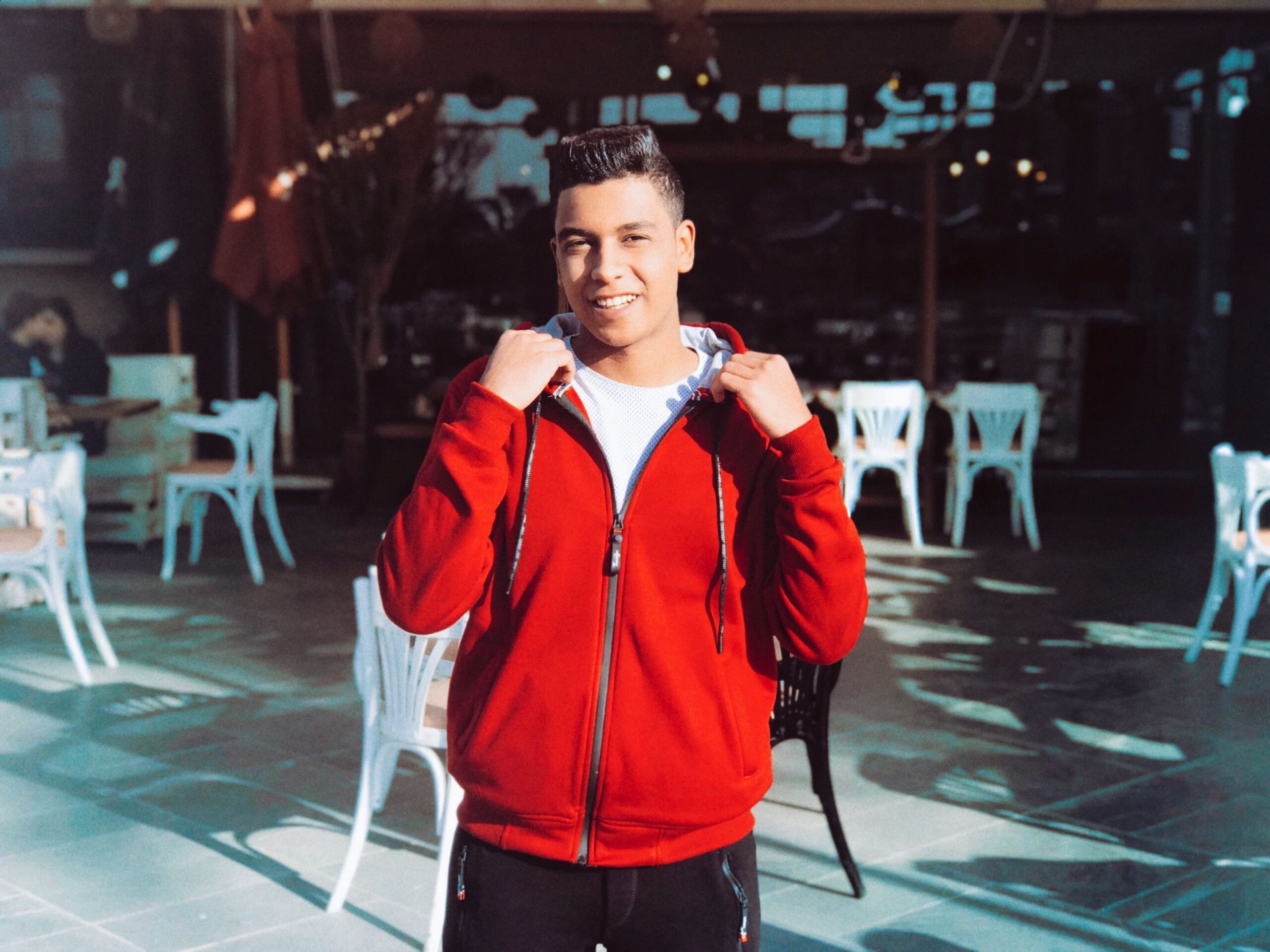 A stylish man in a red hoodie poses confidently in an outdoor setting with white chairs.