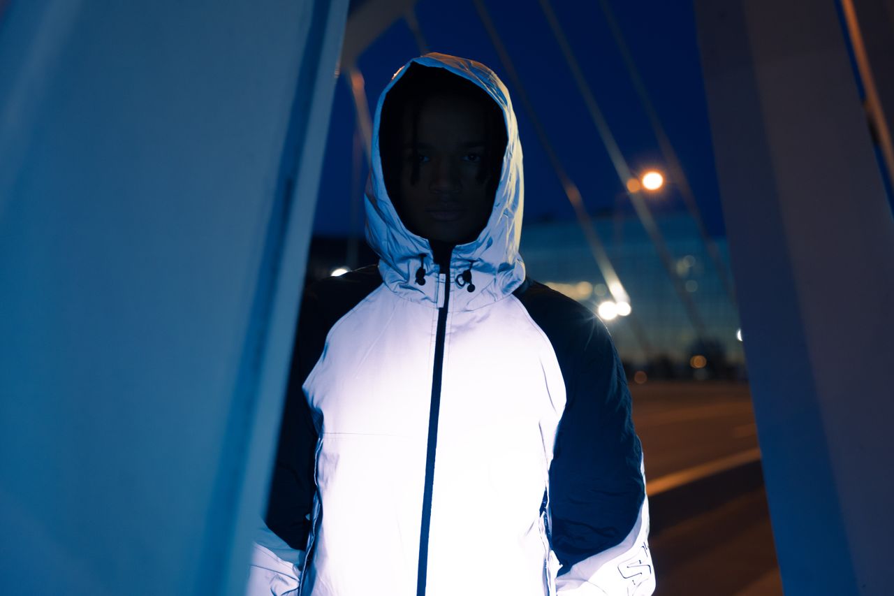 A person in a reflective white hoodie stands on an urban street at night, creating a mysterious atmosphere.