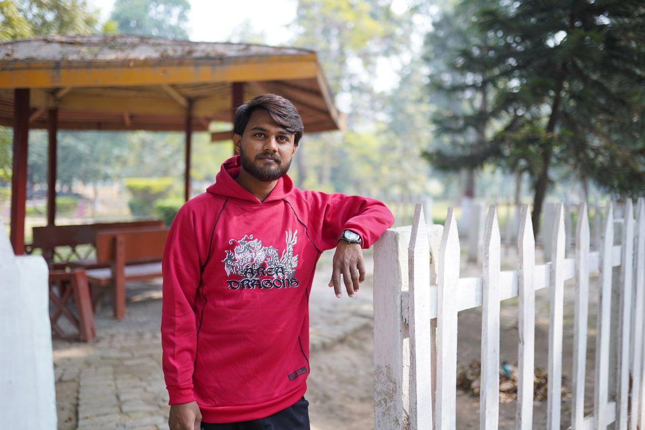 Portrait of a young man with facial hair in a park, wearing a red hoodie and leaning on a white fence.