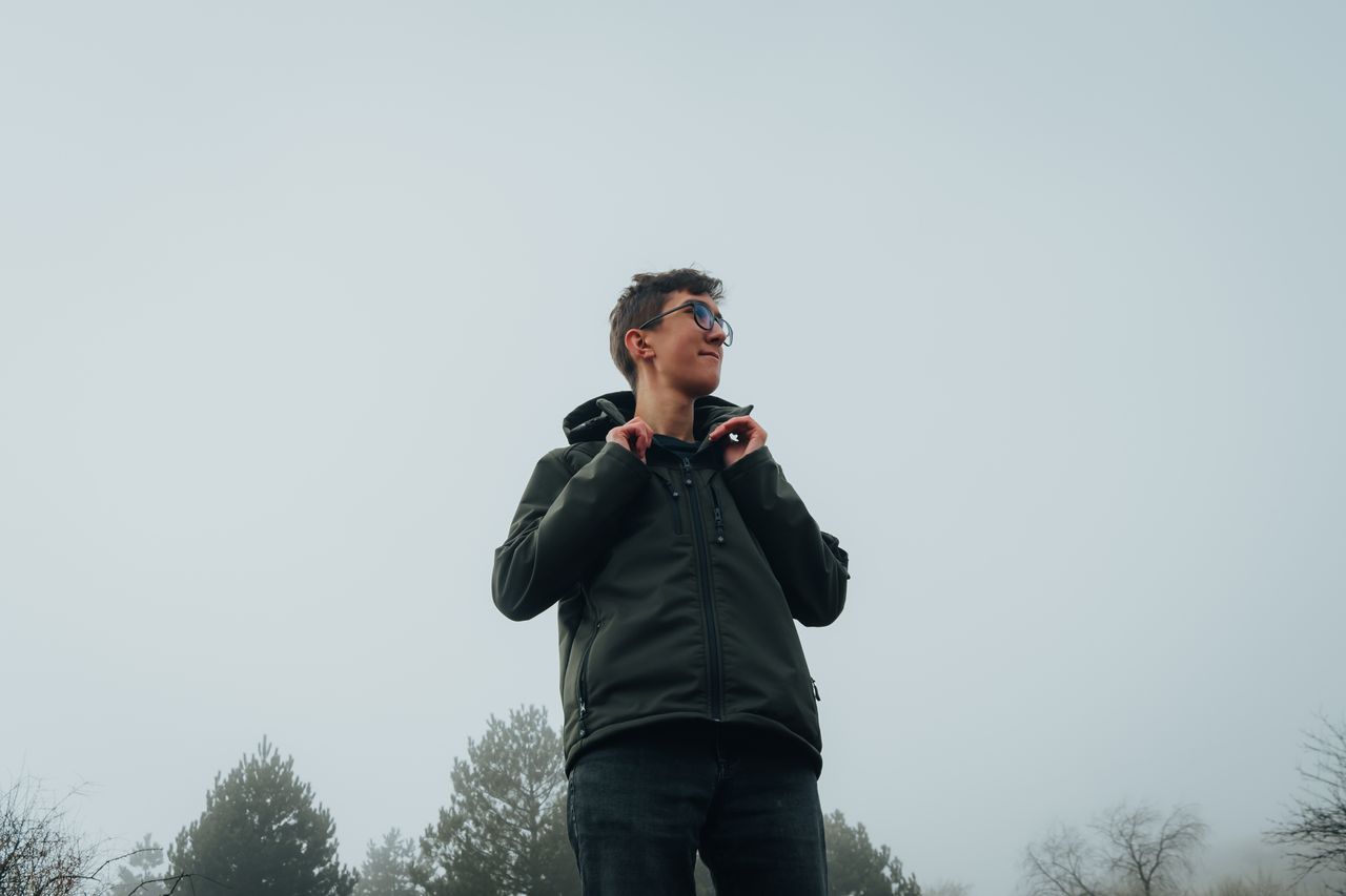 A young man in a jacket stands outdoors amidst foggy trees, creating an adventurous and cool atmosphere.