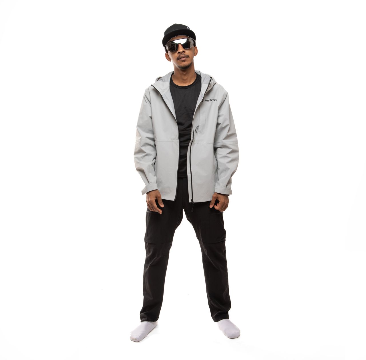 Portrait of a man wearing a cap, sunglasses, and a sporty jacket against a white background.