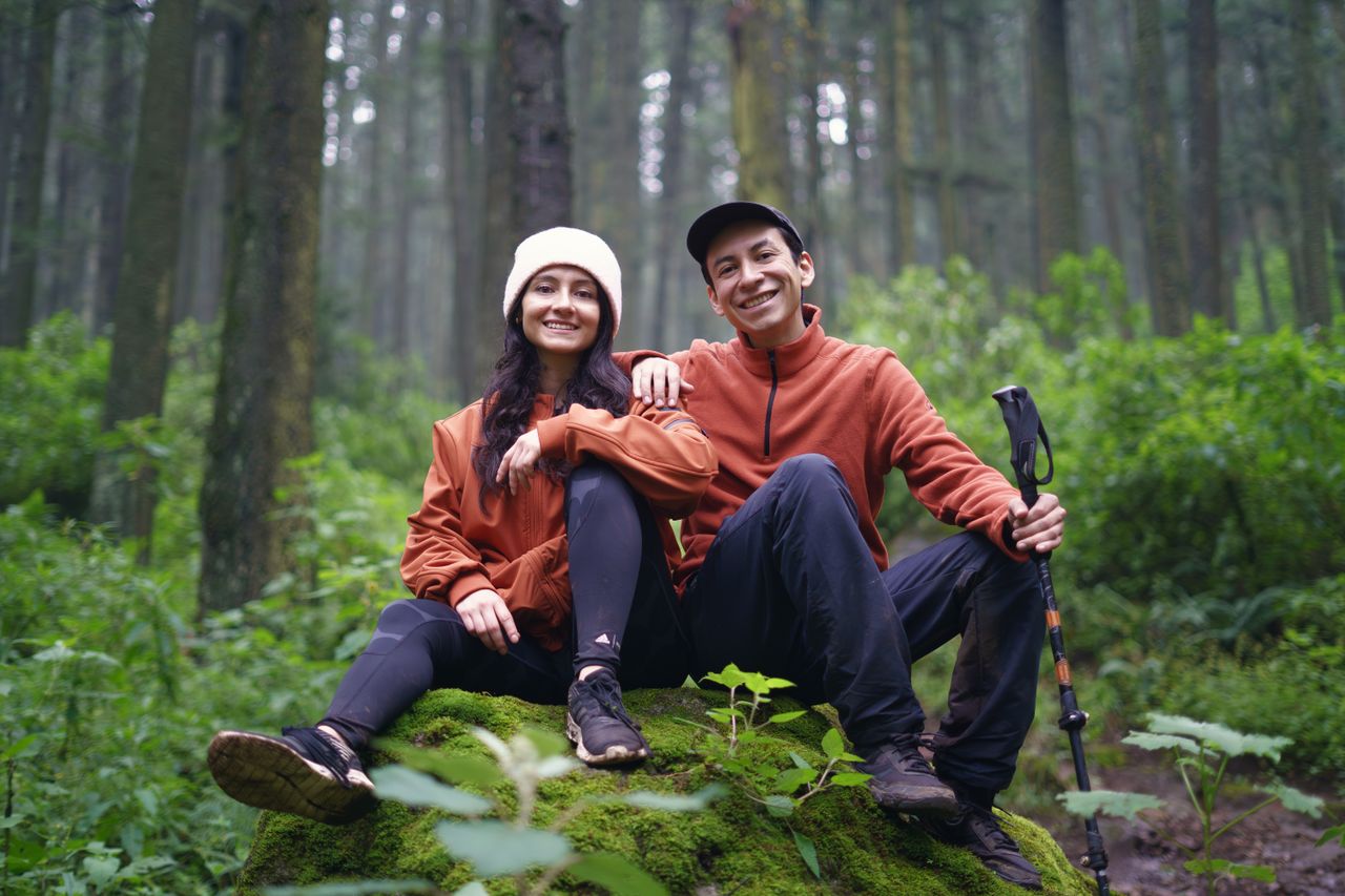 Smiling couple in orange jackets sit on a mossy rock during a forest hike.