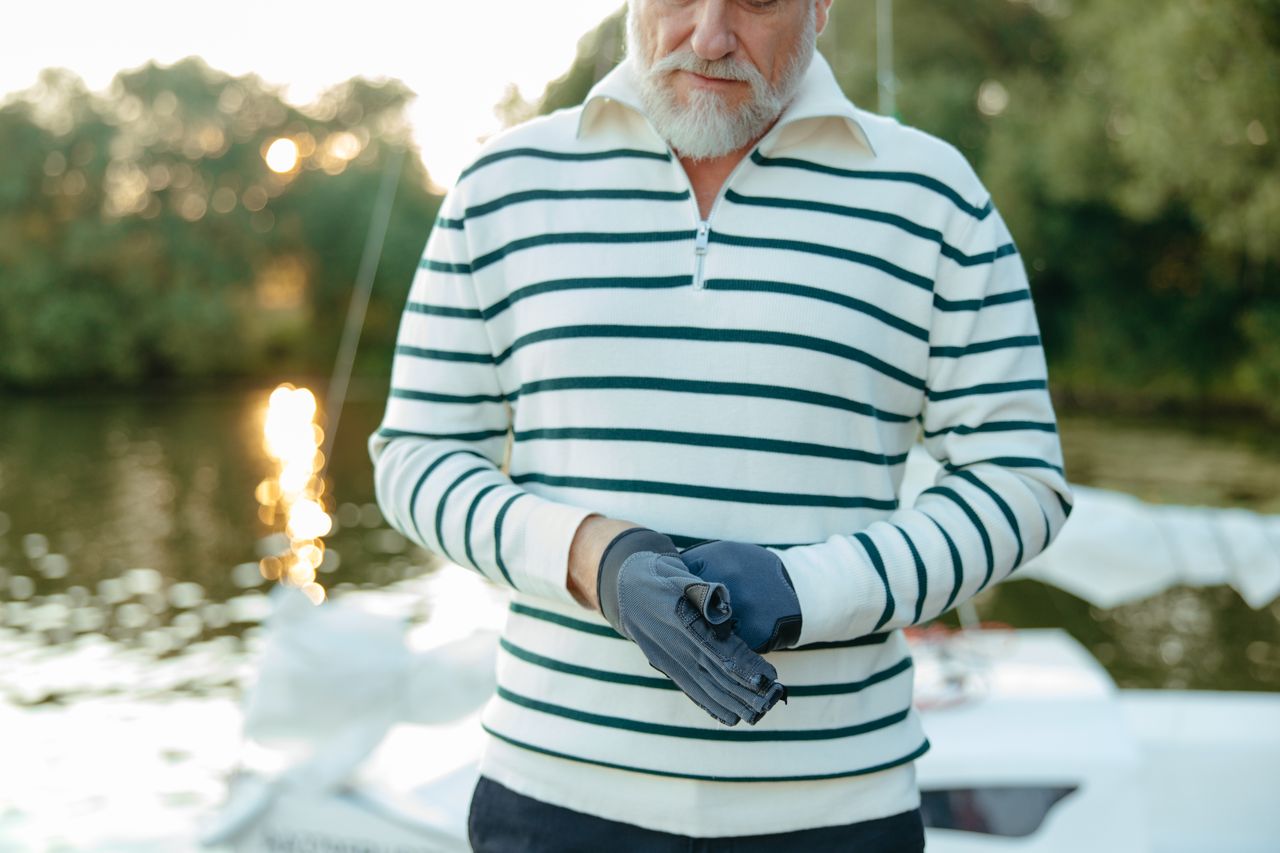 Elderly man wearing gloves by a lake, dressed in a striped pullover, enjoying the outdoors.