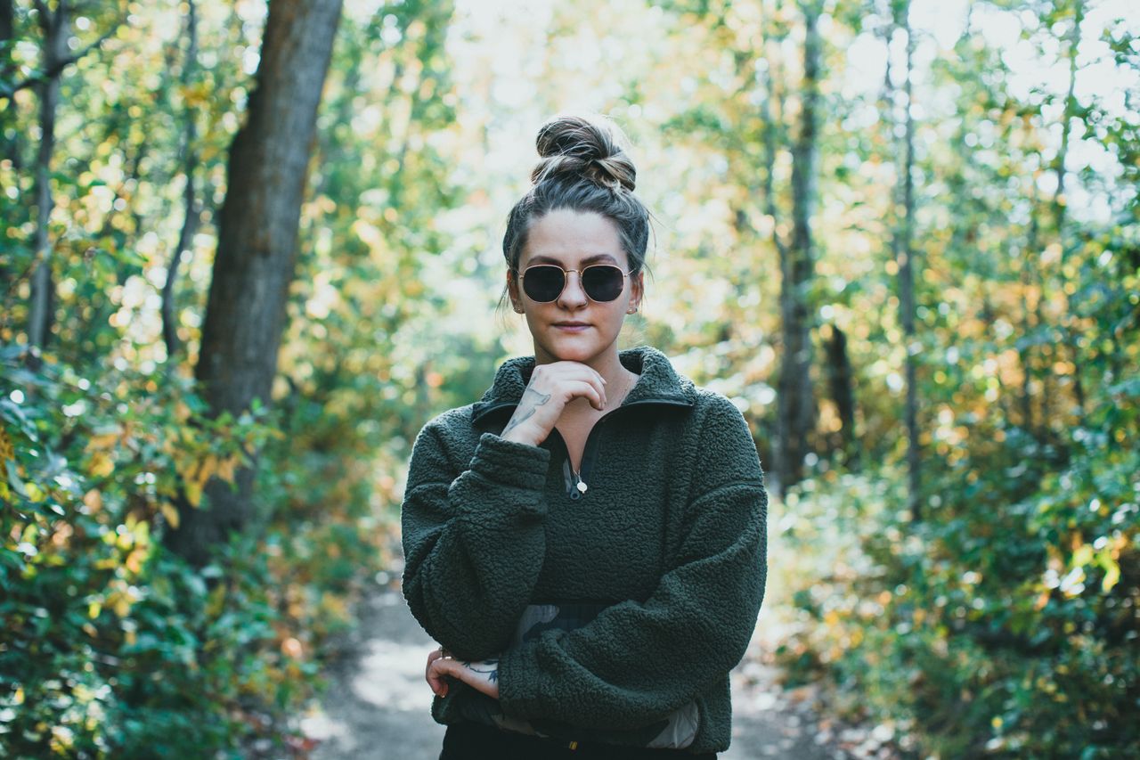 A stylish woman with sunglasses poses confidently in a sun-dappled forest path.
